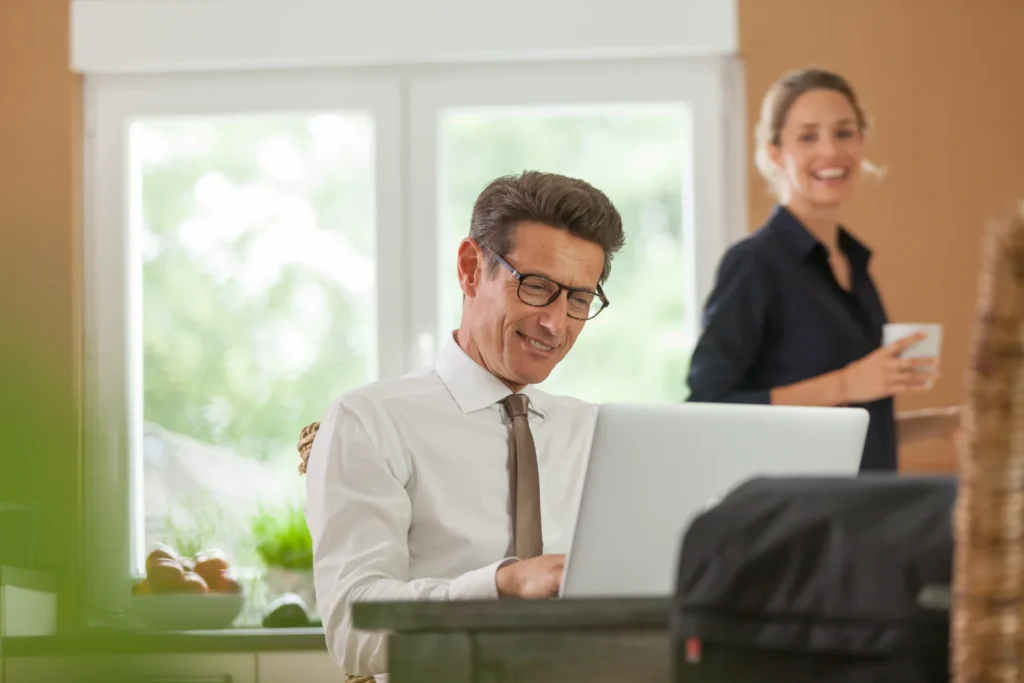 Mann arbeitet lächelnd am Laptop in seiner Küche, im Hintergrund eine Frau mit Kaffeetasse